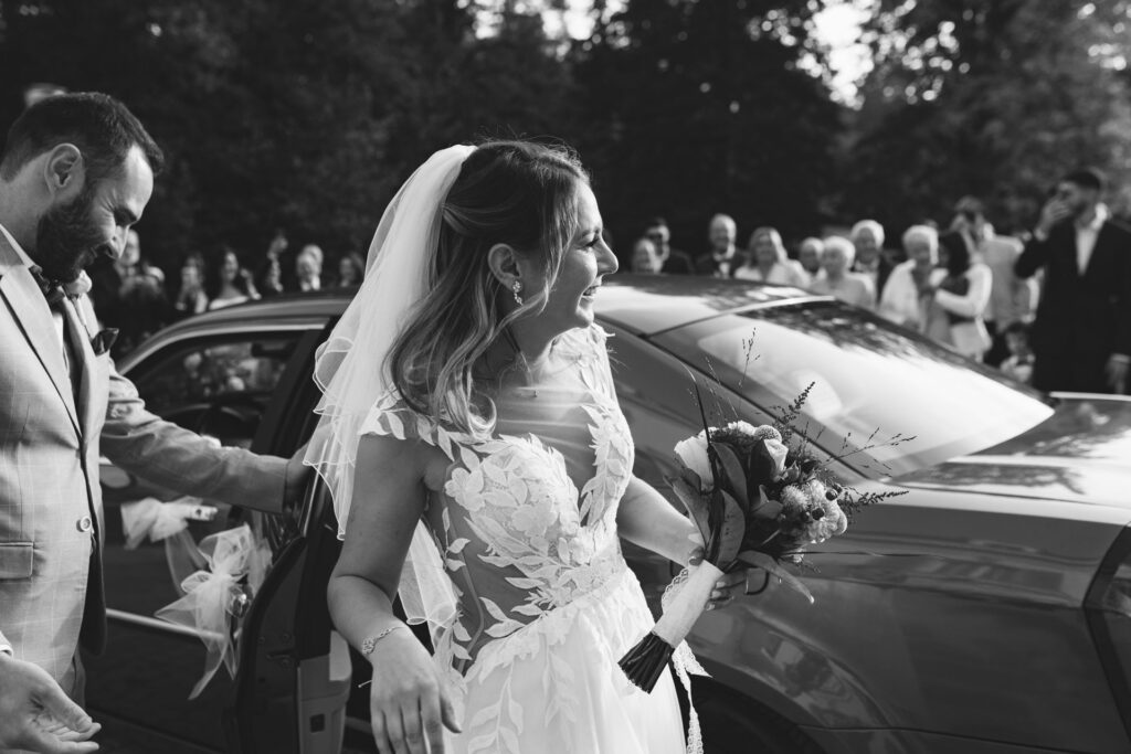 Mariée en robe brodée rejoignant la voiture, entourée de ses invités