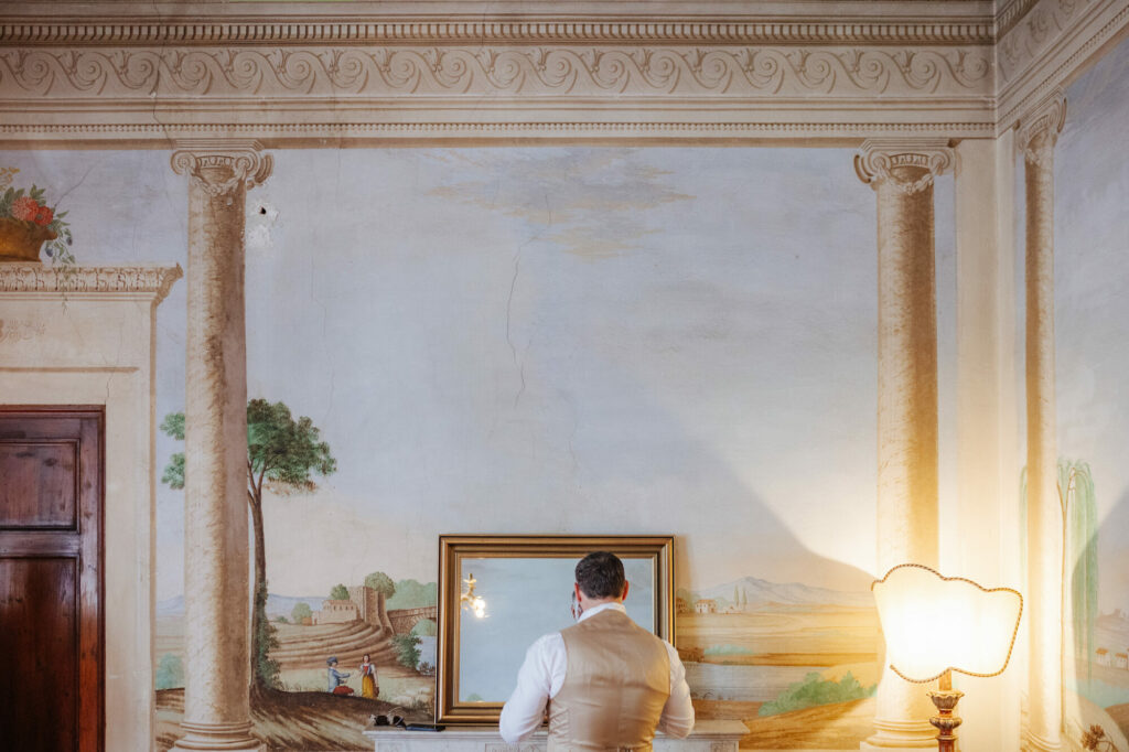 Groom at the mirror with a painted Tuscan landscape behind him